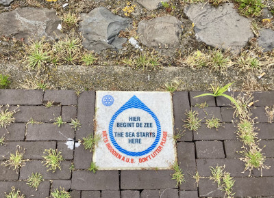 A sign on a brick floor with text inside a blue arrow that reads 'Hier Begint De Zee' / 'The Sea Starts Here'. There are grasses and weeds growing between the paving.